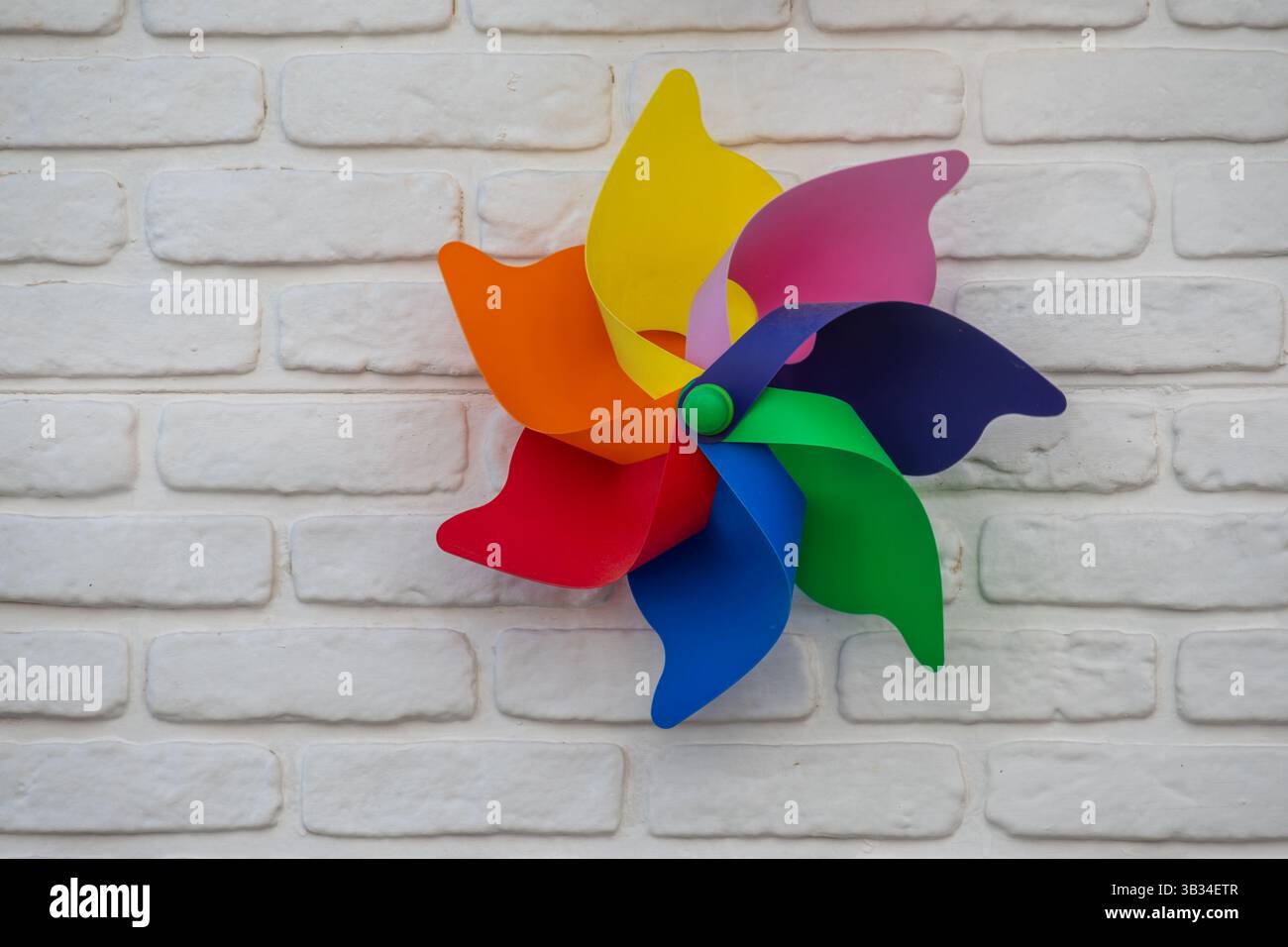 Ein farbenfrohes Spielzeugdrehrad mit sechs leuchtenden Klingen – rot, orange, gelb, rosa, lila, blau und grün – wird an einer weißen Ziegelwand montiert. Stockfoto