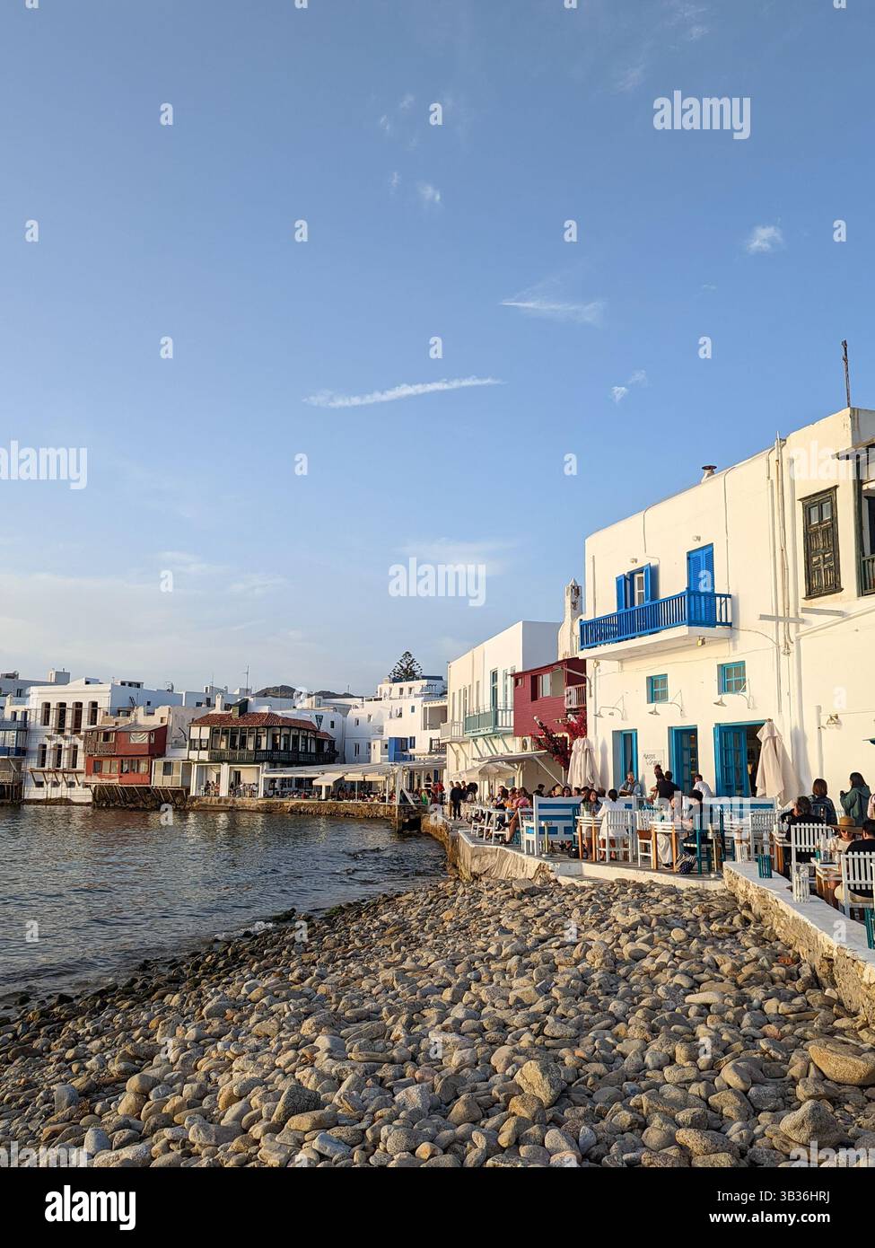 Ein Blick auf Bars und Restaurants an der Küste von Mykonos Stadt, Griechenland, mit Blick auf die Ägäis unter einem blauen Himmel kurz vor Sonnenuntergang - Smartphone-aufgenommenes Stockfoto