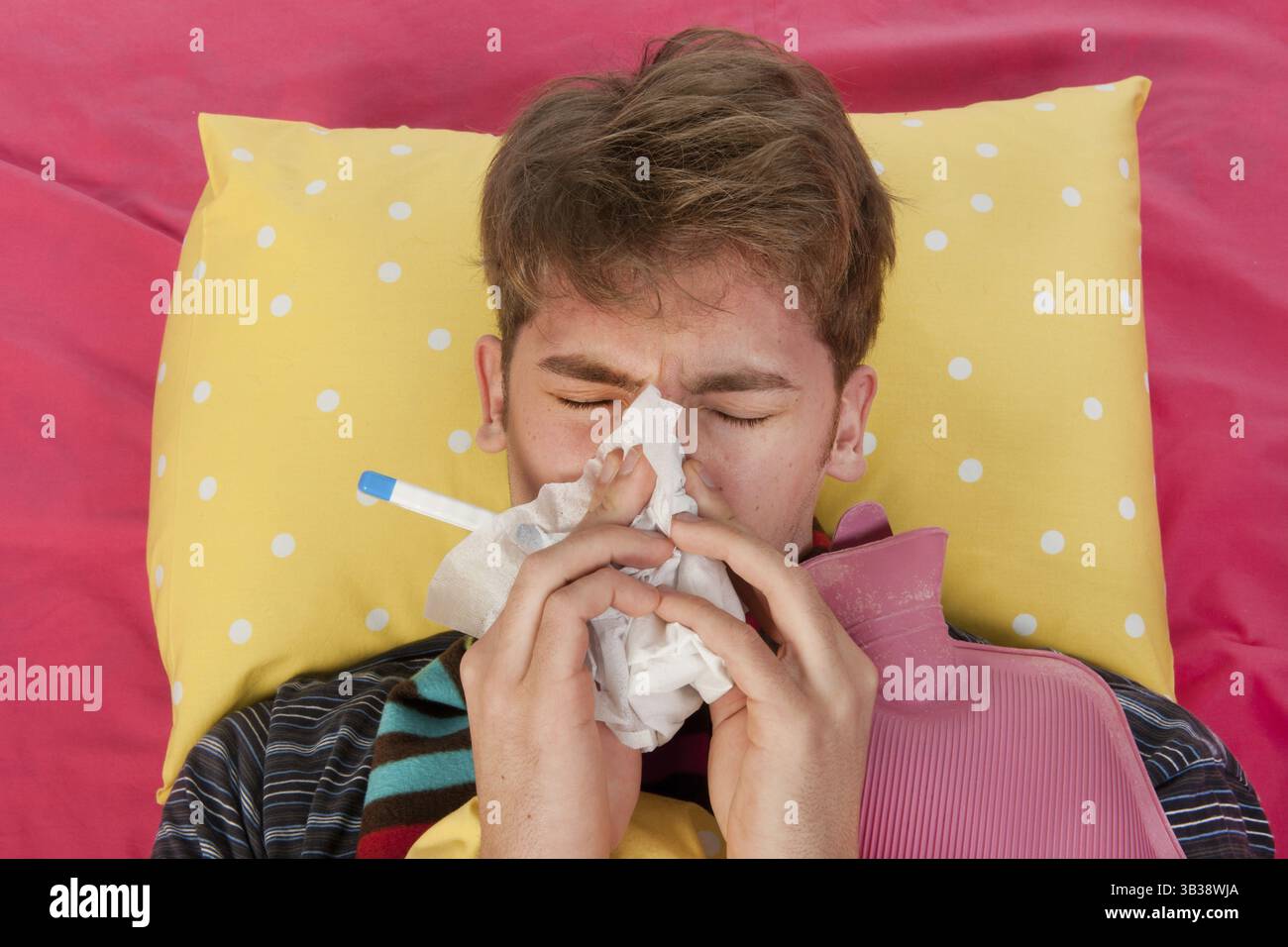 Ein sehr kranker Mann liegt in seinem Bett mit einer warmen Wasserflasche und einem Thermometer, während er sich die Nase bläst Stockfoto