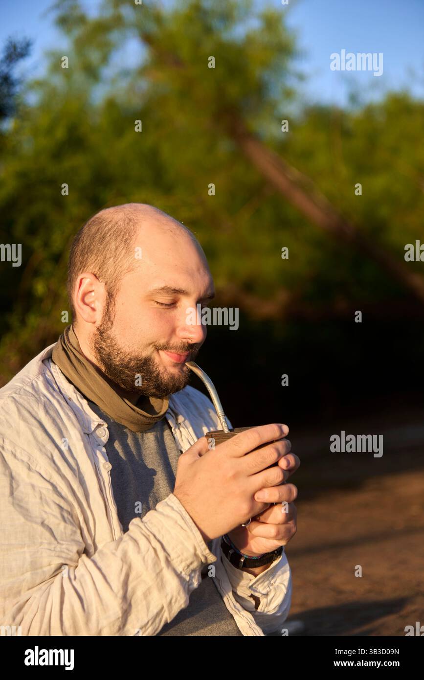 Hispanischer Reisender, der bei Sonnenaufgang am Ufer eines Baches sitzt und die ersten Sonnenstrahlen empfängt, während er Mate trinkt, ein typisches argentinisches Getränk. El Stockfoto