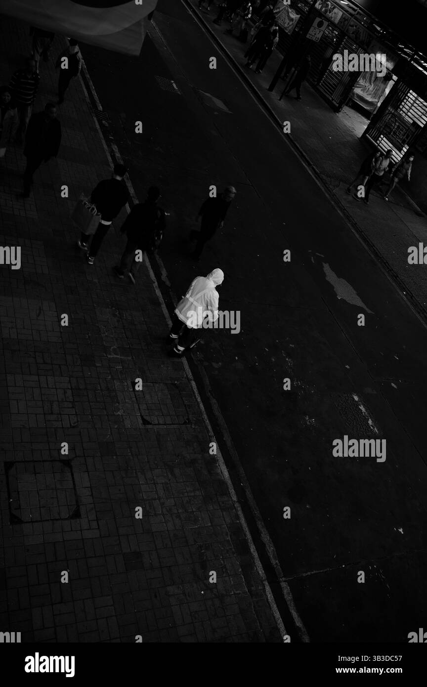 Straßenreiniger in reflektierendem weiß steht leise an der Bordsteinkante, während das Stadtleben in Schwarzweiß fließt Stockfoto