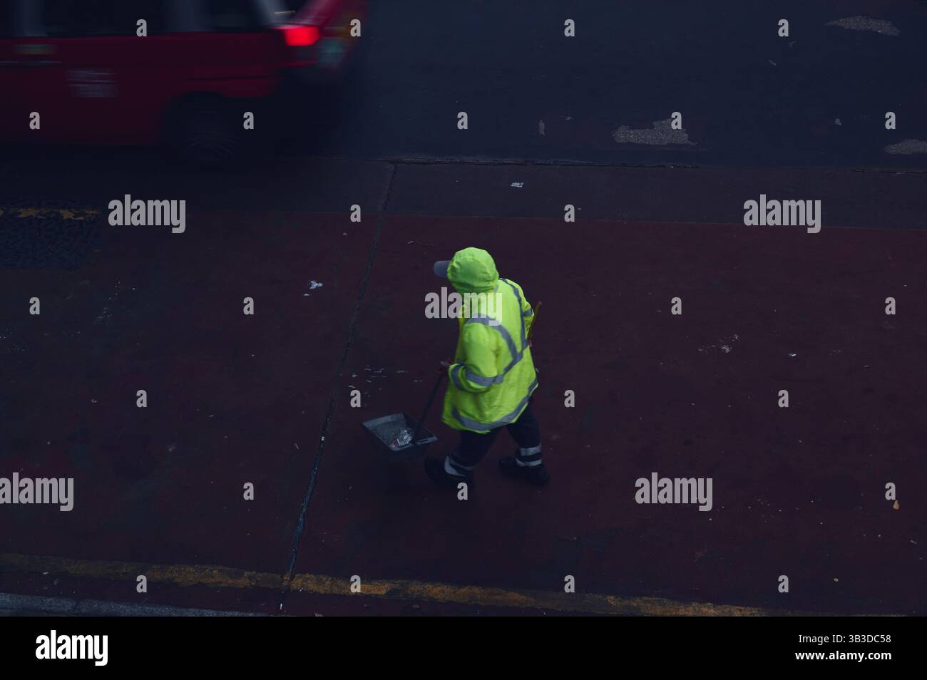 Straßenreiniger in Neonjacke arbeitet fokussiert, während ein rotes Taxi im schwindenden Licht der Stadt vorbeirauscht Stockfoto