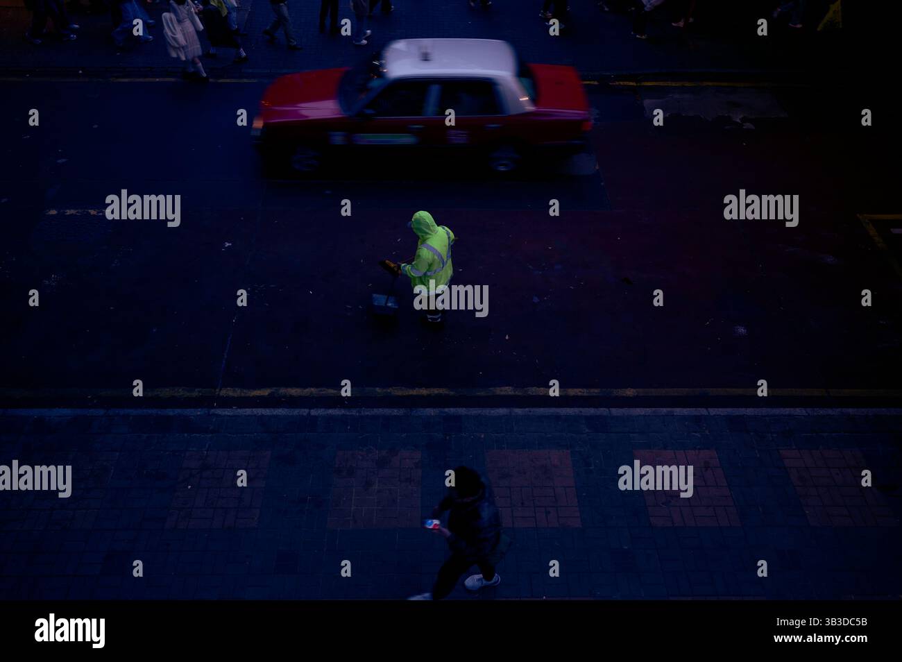 Straßenreiniger in Neon steht still, während ein rotes Taxi unter Abendlichtern vorbeifährt Stockfoto