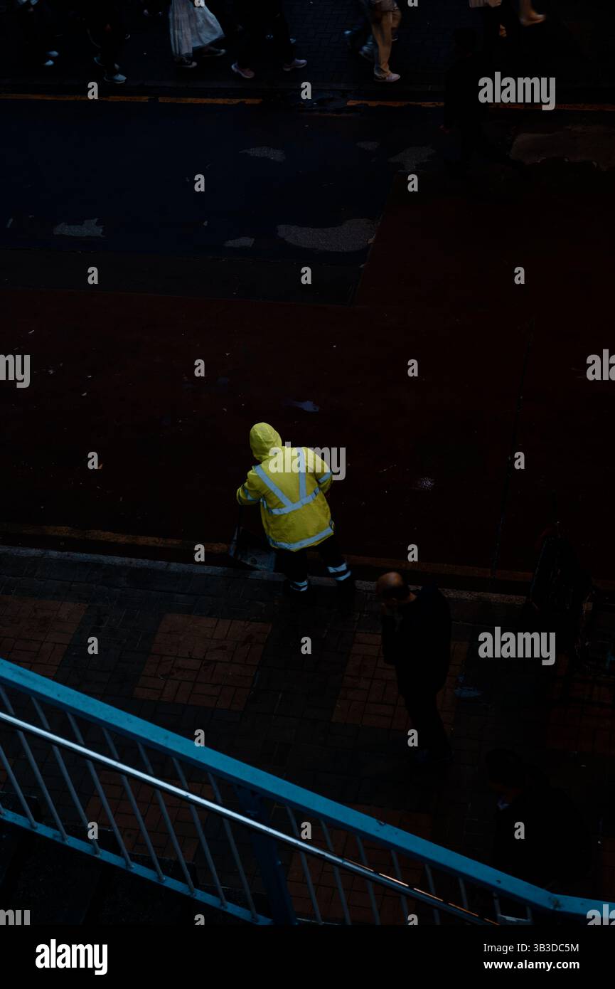 Ein Stadtarbeiter in Neonausrüstung fliegt unbemerkt, wenn die Menschenmassen am Abend auf einer dunklen Stadtstraße vorbeiziehen Stockfoto