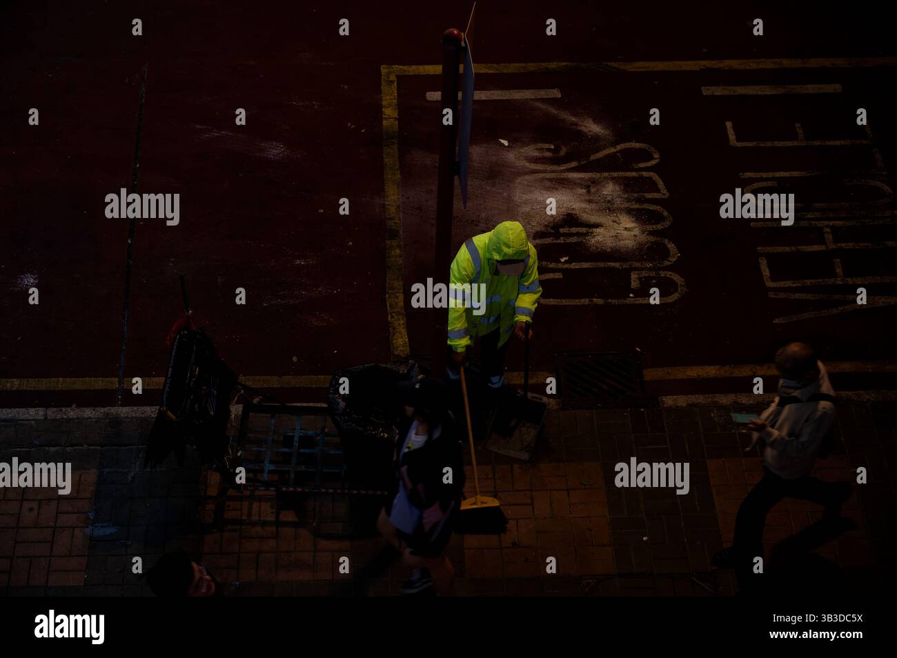 Straßenreiniger in Neonjacke fegt leise unter die Lichter der Stadt, während Fußgänger in der Nacht vorbeikommen Stockfoto