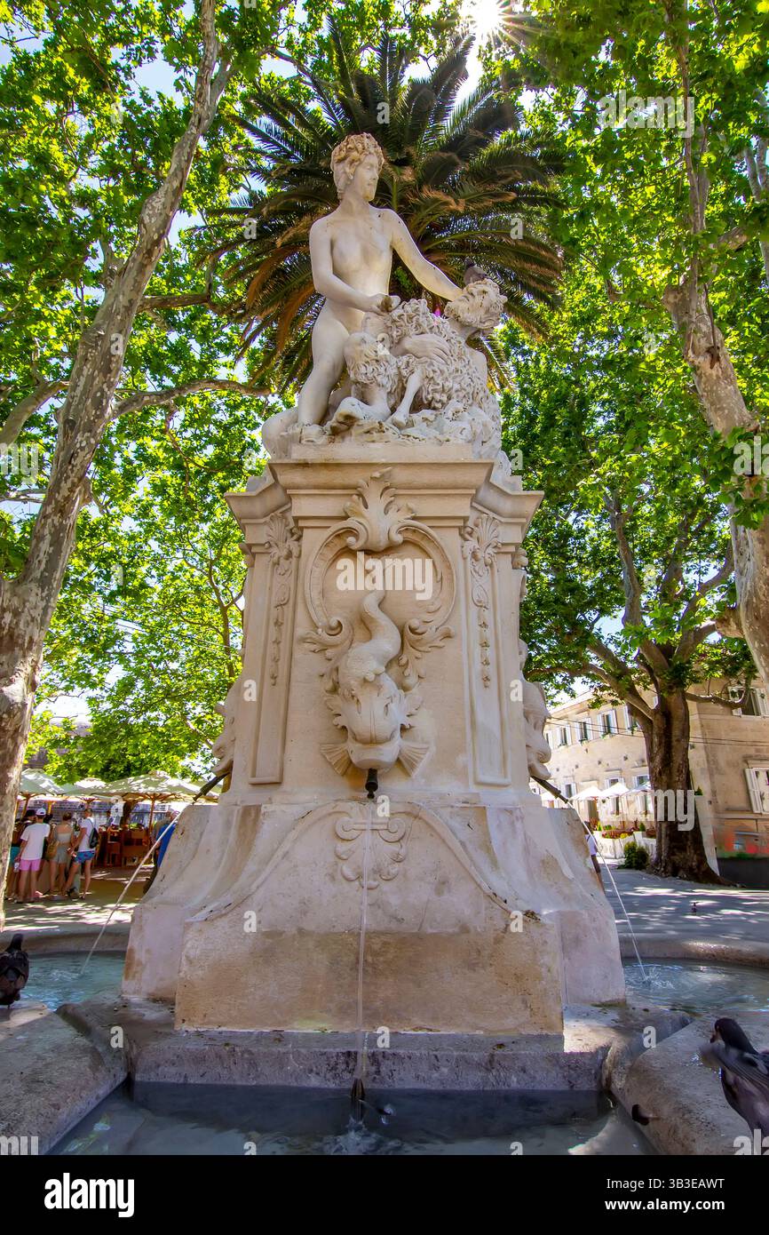 Eine barocke Skulptur eines Satyrn und einer Nymphe krönt den Amerling-Brunnen in Dubrovnik, umgeben von üppigen Bäumen und Stadtleben Stockfoto