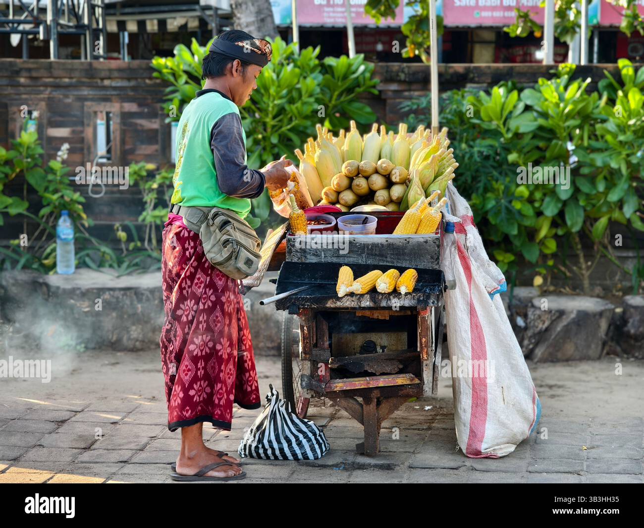 Seminyak, Bali, Indonesien - 29. April 2025 : Ein örtlicher balinesischer Maishändler grillt Mais auf einem Holzkohleofen Stockfoto