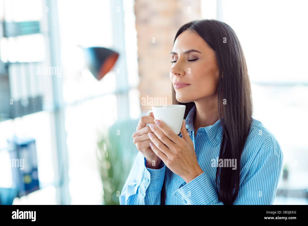 Junge Geschäftsfrau, die einen Moment der Entspannung genießt, während sie Kaffee in einer hellen und modernen Büroumgebung schlürft Stockfoto