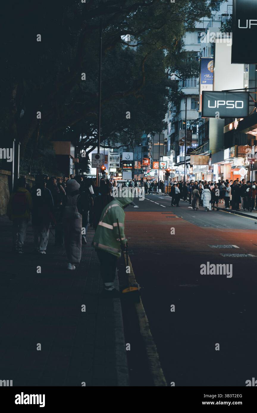 Straßenreiniger in Neonausrüstung fegt leise einen Bordstein in Hongkong, während Menschenmassen unter dem Glanz der Stadt spazieren gehen Stockfoto