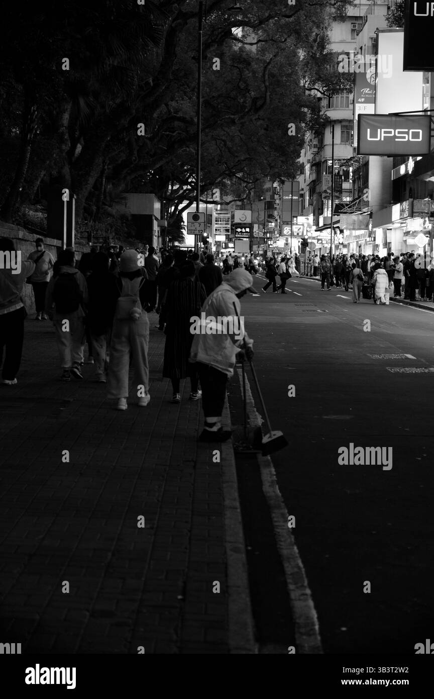 Straßenreiniger in reflektierender Ausrüstung pflegt leise die Bordsteinkante, während sich die Menschenmassen durch eine geschäftige Nacht bewegen Stockfoto