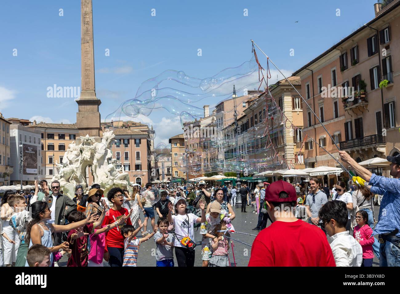 Rom, Italien - 23. April 2025: Eine lebhafte Menschenmenge versammelt sich auf einem belebten Stadtplatz, während Kinder die riesigen Blasen jagen. Die Sonne scheint hell Stockfoto