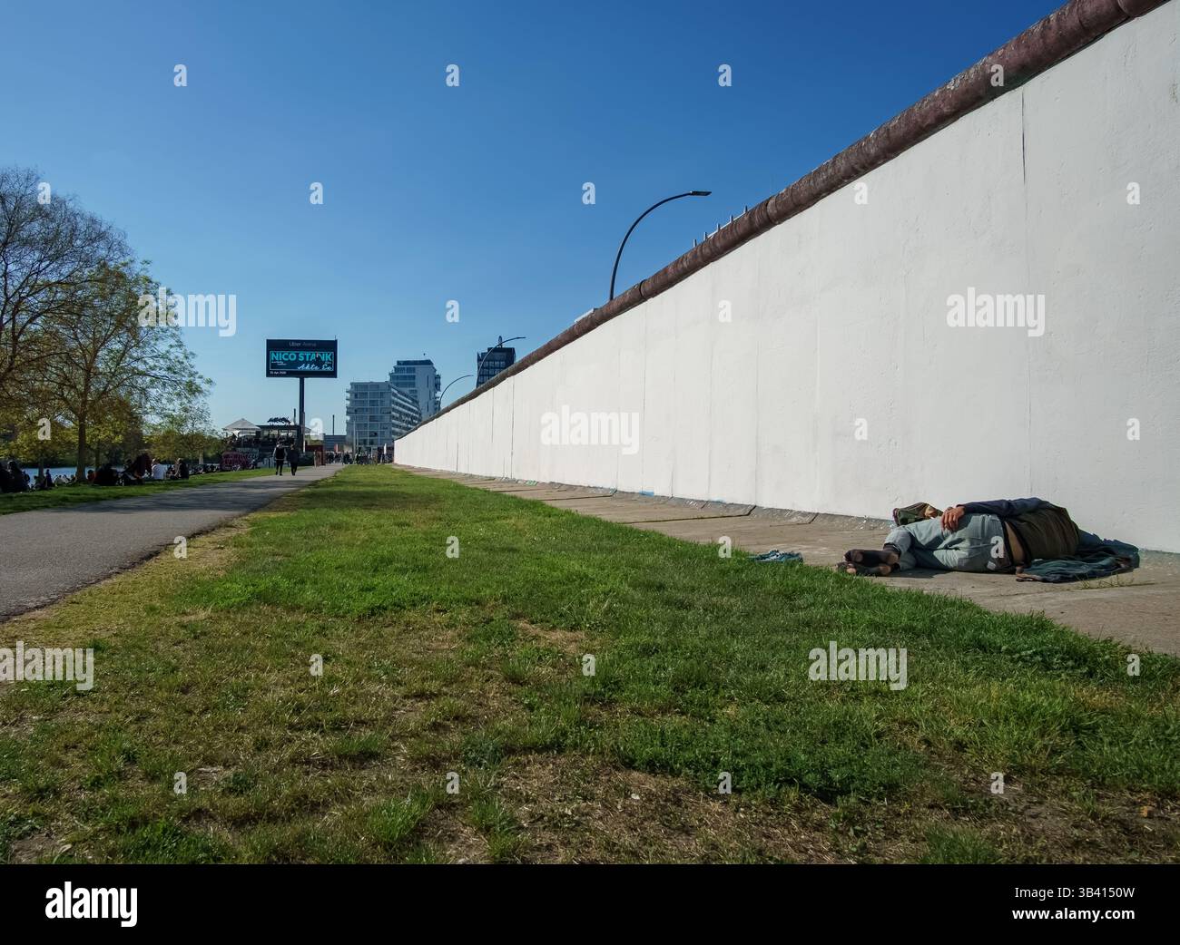 Obdachloser Mann, der vor der vor kurzem geweihten Berliner Mauer in der East Side Gallery liegt und die städtische Realität und soziale Herausforderungen widerspiegelt. Stockfoto