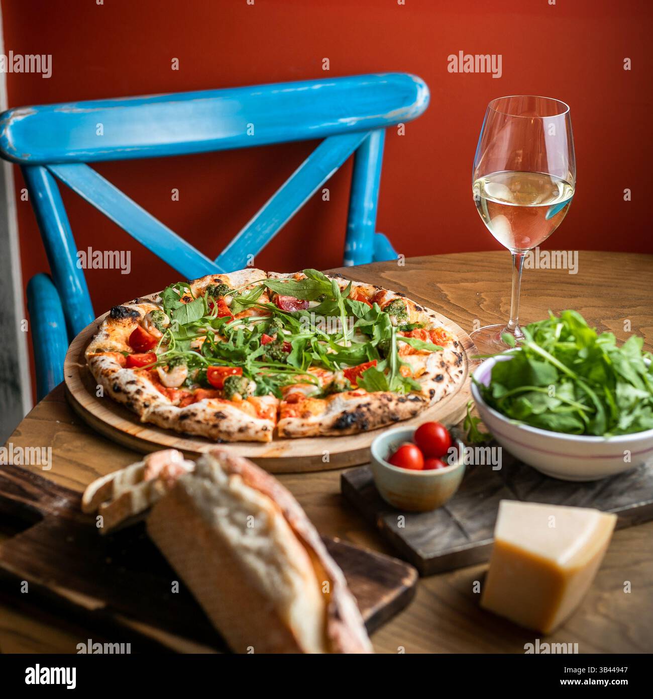 Seitlicher Blick auf Pizza auf Holzbrett, ein Glas Weißwein, Brotscheiben und Käse, Grün und Tomaten in einer kleinen weißen Schüssel Stockfoto