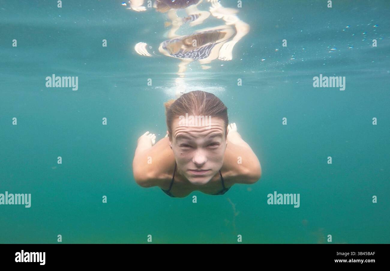 Junge Frau schwimmt unter dem Wasser in Richtung Lanze und blickt in die Lanze. Realistischer Gesichtsausdruck unter Wasser durch Atemanhalten. Stockfoto
