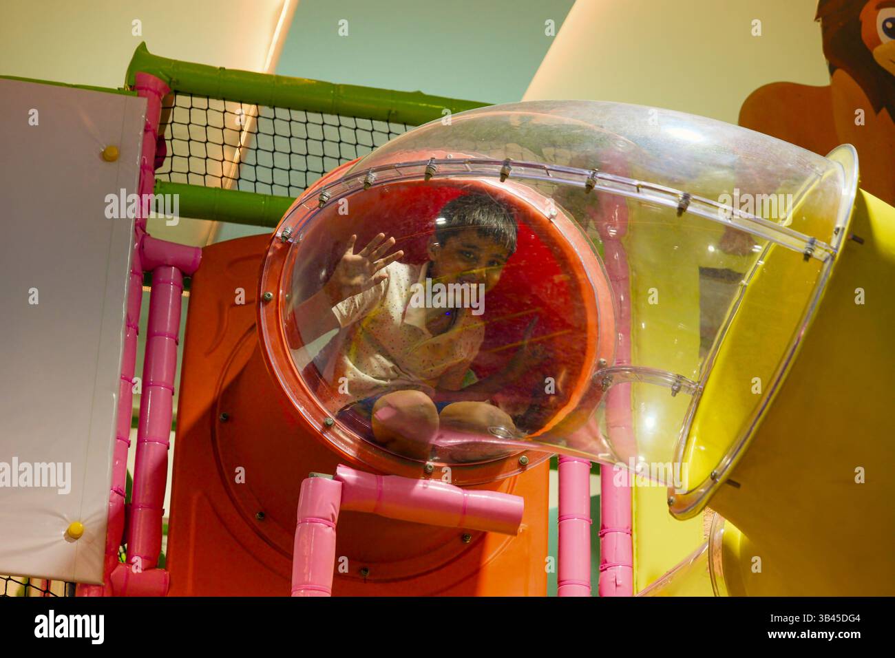 Ein Junge winkt und lächelt aus einem bunten Spieltunnel auf einem Spielplatz. Stockfoto