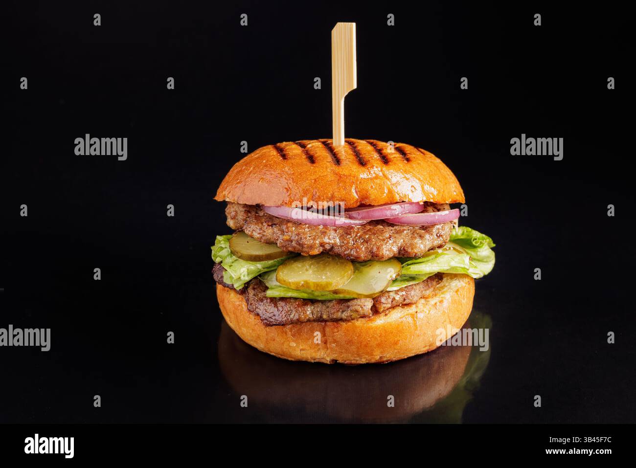 Saftiger gegrillter Rindfleischburger mit Gurken, Salat, Zwiebeln in einem gerösteten Brötchen auf schwarzem Hintergrund, Nahaufnahme. Stockfoto