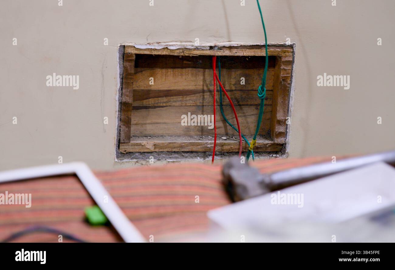 Elektriker, der während des Renovierungsprozesses an freiliegenden Kabeln in einer Wand arbeitet. Werkzeuge und Materialien sind in der Nähe verstreut. Stockfoto