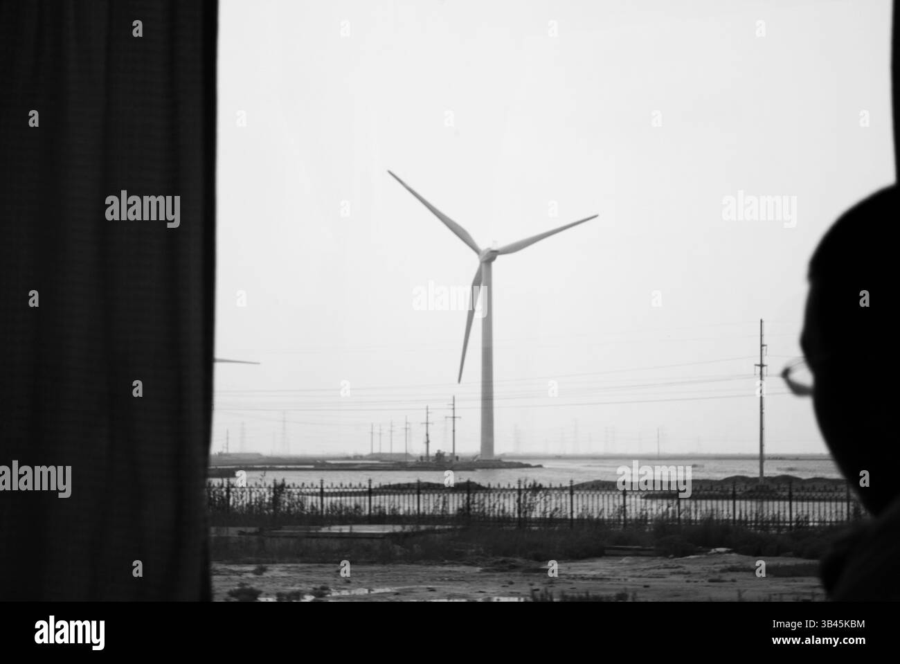 Windkraftanlage durch ein Fenster gesehen, stehen Schwarzweißbild / Windkraftanlagen hoch vor einem grauen Himmel, während ein Beobachter von drinnen blickt und das Wesen erneuerbarer Energien in einer Industrielandschaft erfasst Stockfoto