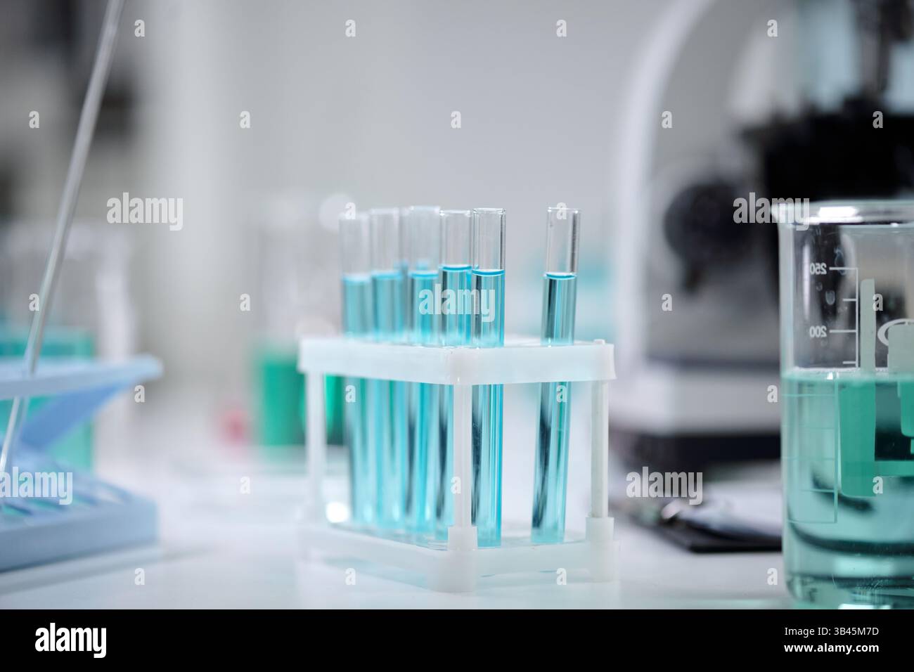 Laborausrüstung mit mehreren Reagenzgläsern, die mit einer leuchtend blauen Flüssigkeit gefüllt sind. Im Hintergrund ist ein Becher mit einer Pipette zu sehen, was auf eine wissenschaftliche Versuchsumgebung hinweist Stockfoto