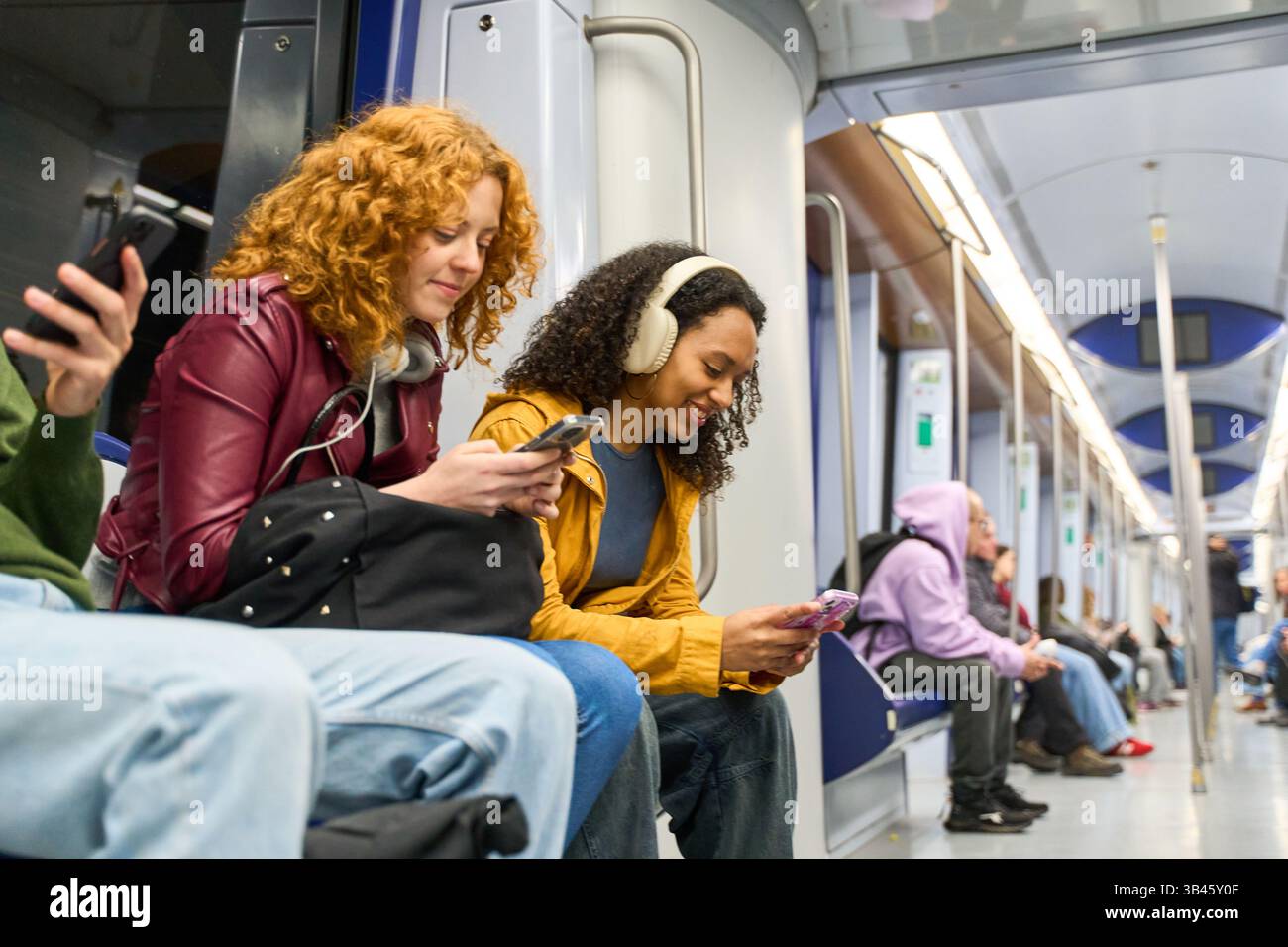 Zwei junge Frauen sitzen in einer U-Bahn, benutzen ihre Smartphones und hören Musik über Kopfhörer während ihrer täglichen Fahrt Stockfoto