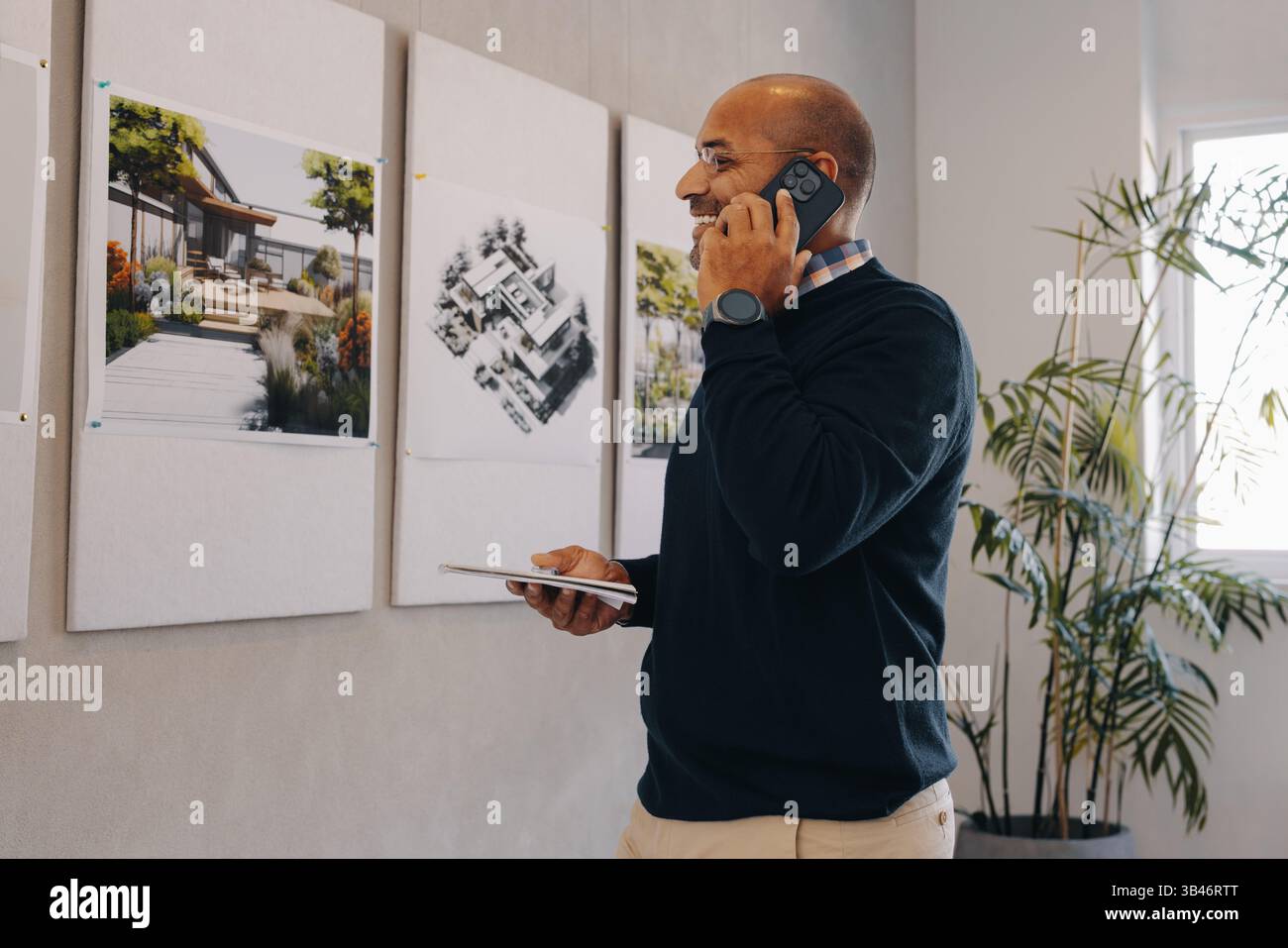 Ein engagierter Projektmanager spricht am Telefon, während er die an einer Wand befestigten Entwurfspläne untersucht. Die Einstellung stellt eine professionelle Umgebung mit einem f dar Stockfoto