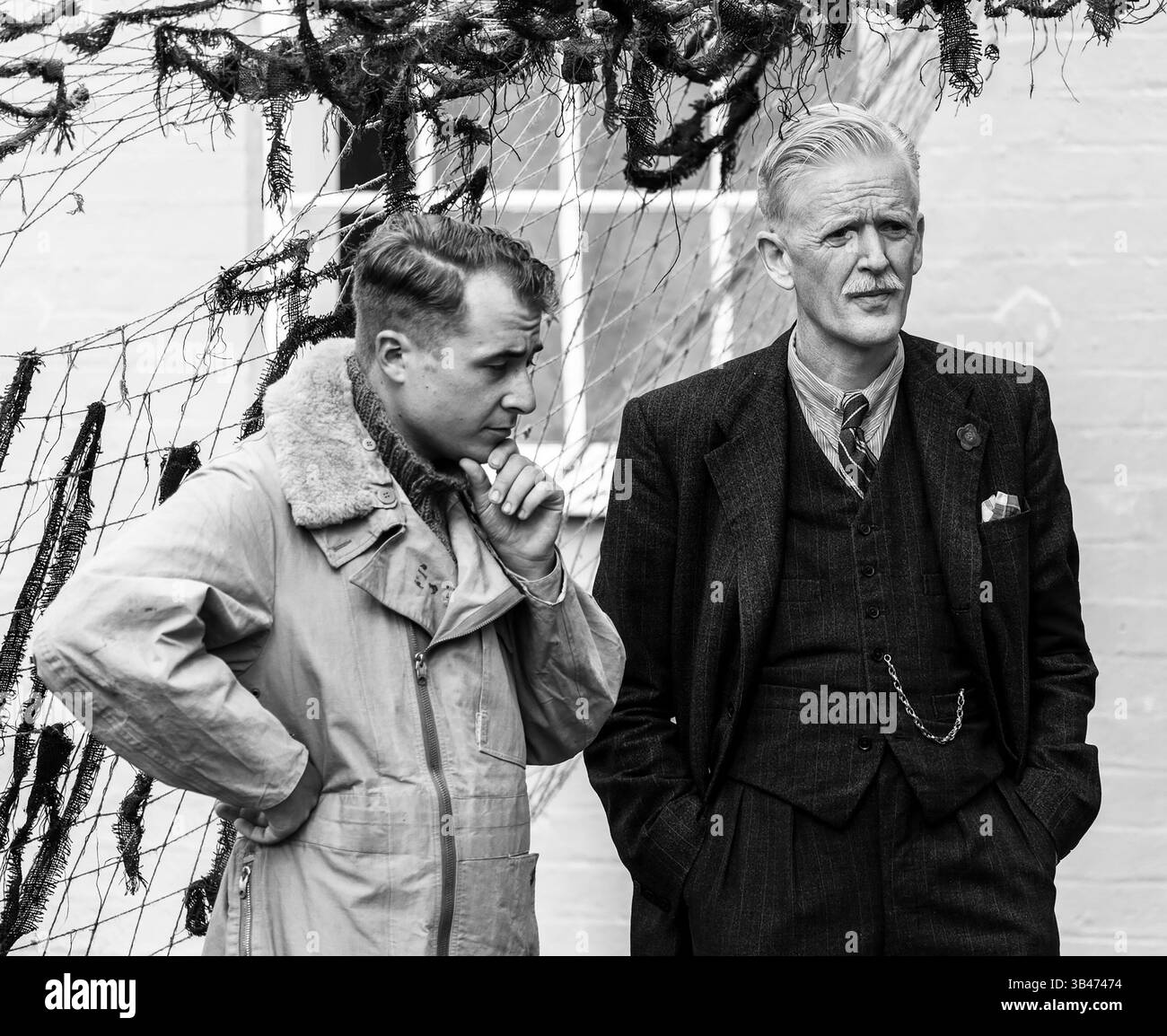 Zwei Männer, einer in einer Pilotenjacke und der andere in einem Anzug, stehen in einer nachdenklichen Unterhaltung vor dem Hintergrund des Netzes in Duxford. Stockfoto