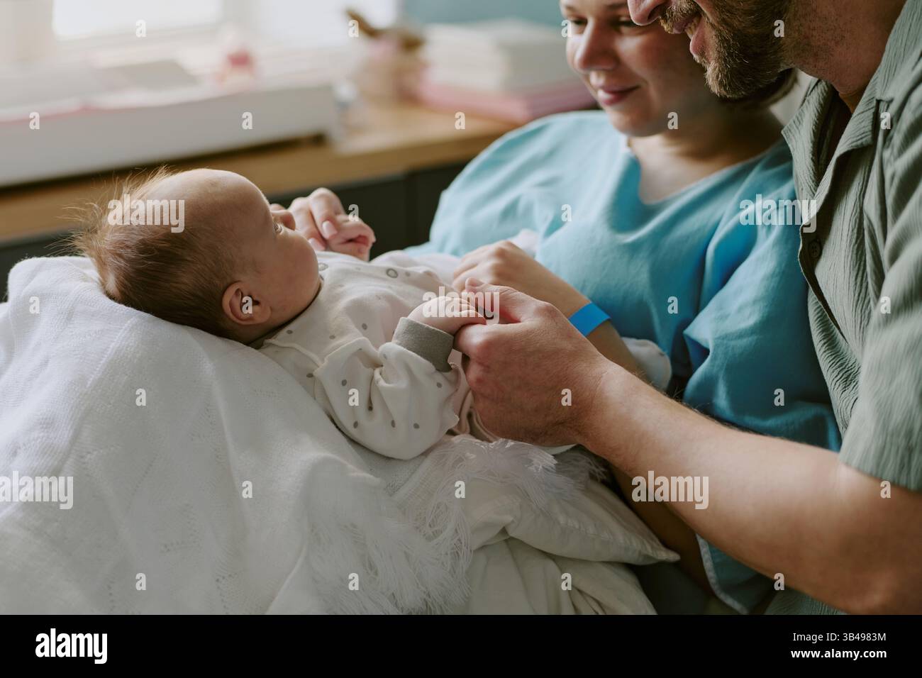 Eltern halten das Neugeborene in weiße Decke gewickelt, während sie einen zärtlichen Moment teilen. Familie genießt enge und intime Interaktion in der Nähe des Fensters Stockfoto