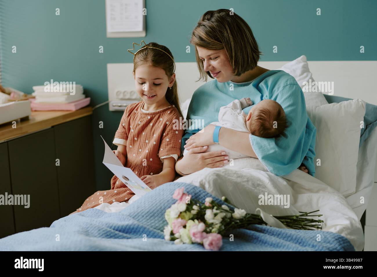 Mutter sitzt im Krankenhausbett, hält Neugeborene fest, während Tochter mit einer Karte in der Nähe sitzt. Ein Zimmer voller Glück, Blumen und einer blauen Decke Stockfoto