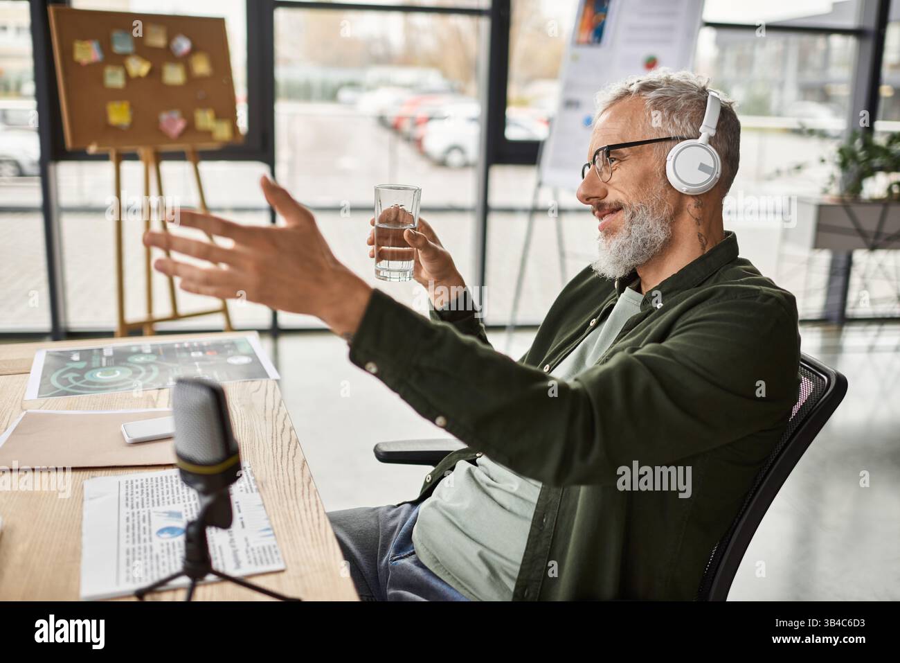 Ein Mann mit Kopfhörern genießt ein Gespräch, während er mit einem Glas Wasser an einem Schreibtisch sitzt. Stockfoto