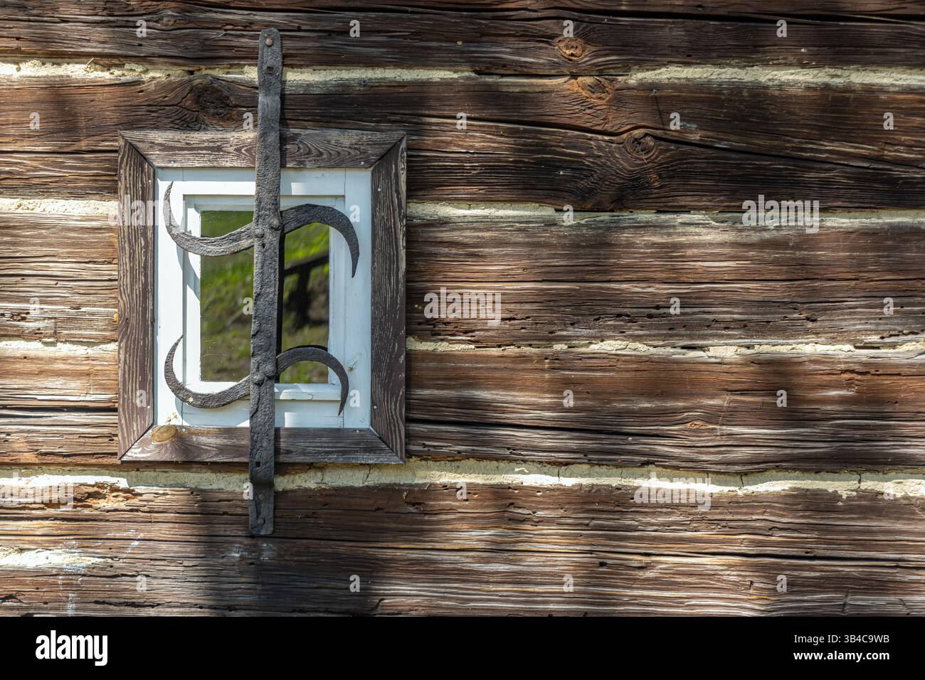 Kleines Fenster an der Wand eines alten Holzhauses mit einem schmiedeeisernen Grill. Stockfoto