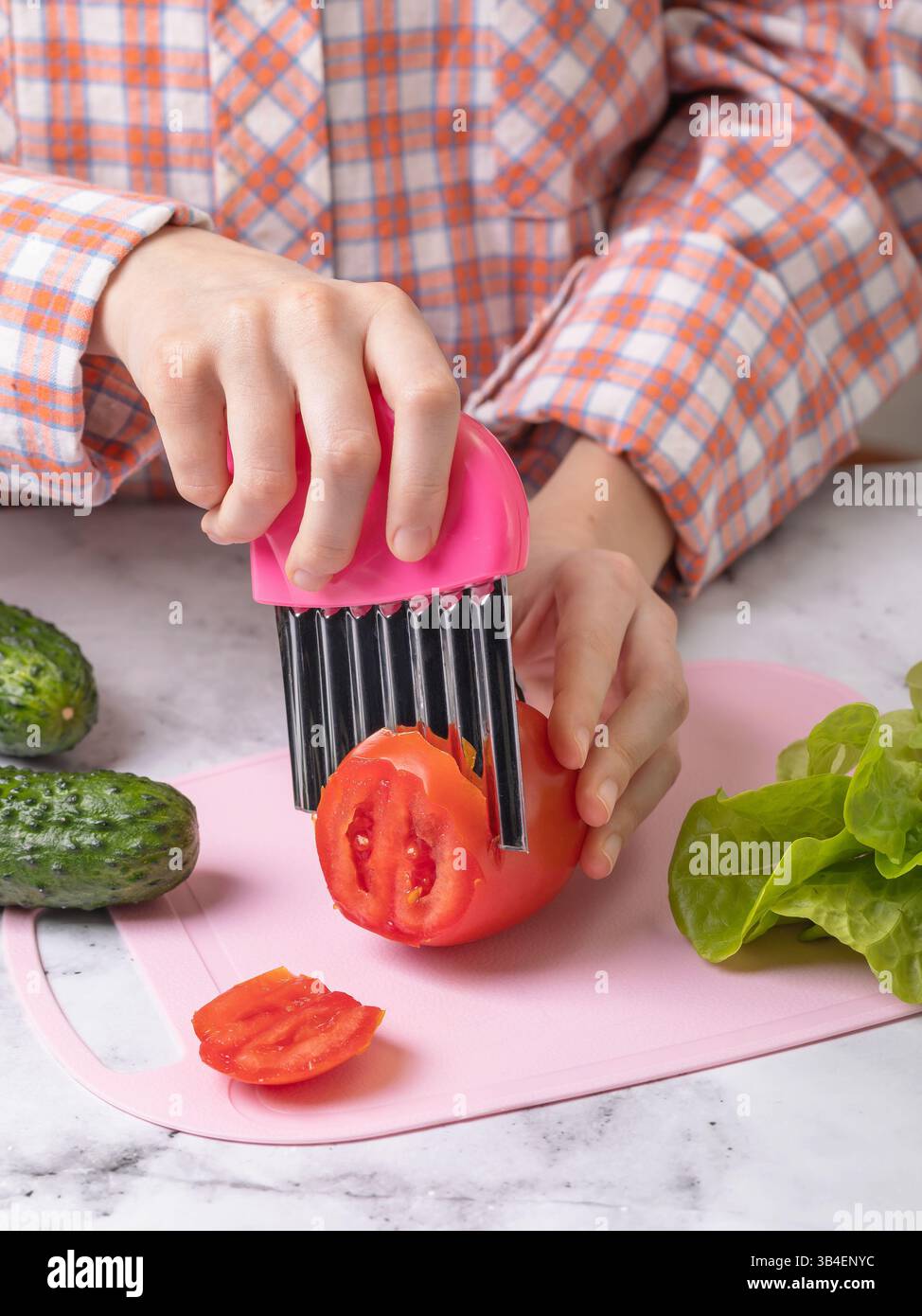 Kind, das mit der Zubereitung von Salat und dem Schneiden von Tomaten mit einem welligen Edelstahlmesser auf rosafarbenem Schneidebrett beschäftigt ist Stockfoto