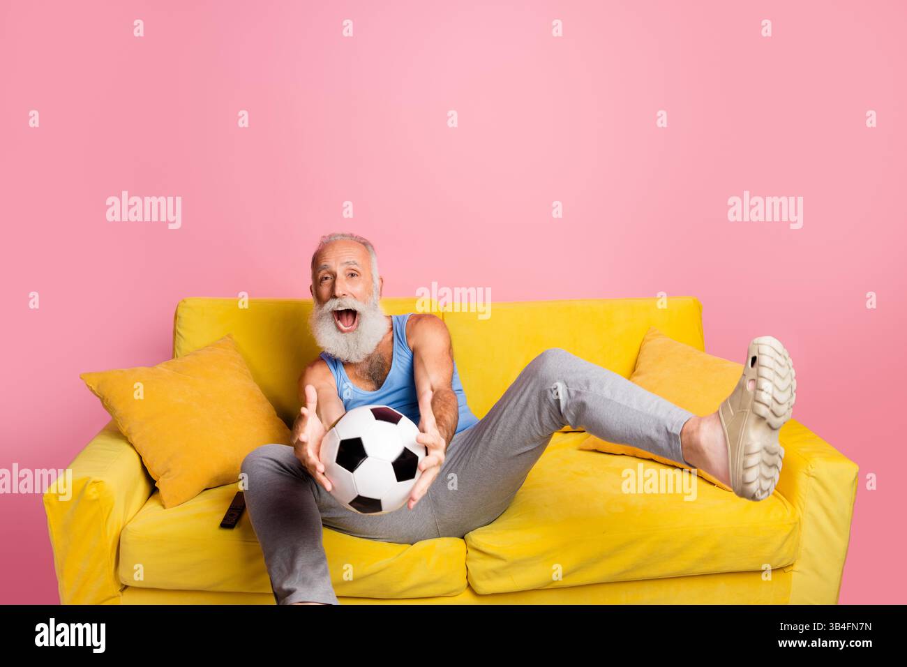 Fröhlicher älterer Mann mit Bart, der Freizeit auf einem lebhaften gelben Sofa vor rosafarbenem Hintergrund genießt Stockfoto