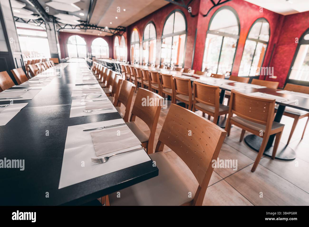 Lange Reihe von Holzstühlen und ordentlich angeordnete Plätze mit Besteck auf schwarzen Tischen in einem hellen, eleganten Restaurant mit Bogenfenstern und Stockfoto