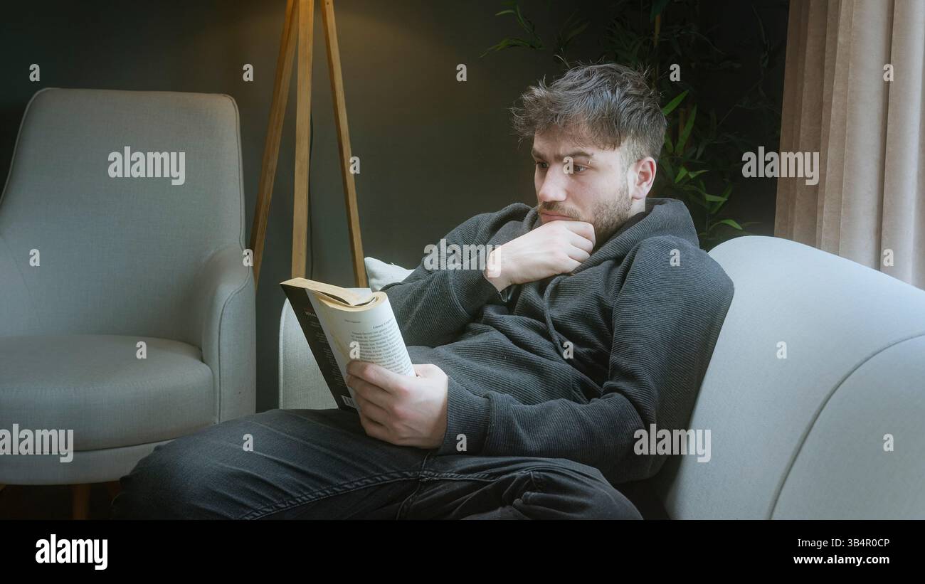 Junger Mann liest ein Buch und entspannt sich in einem gemütlichen Wohnzimmer mit warmer Beleuchtung Stockfoto