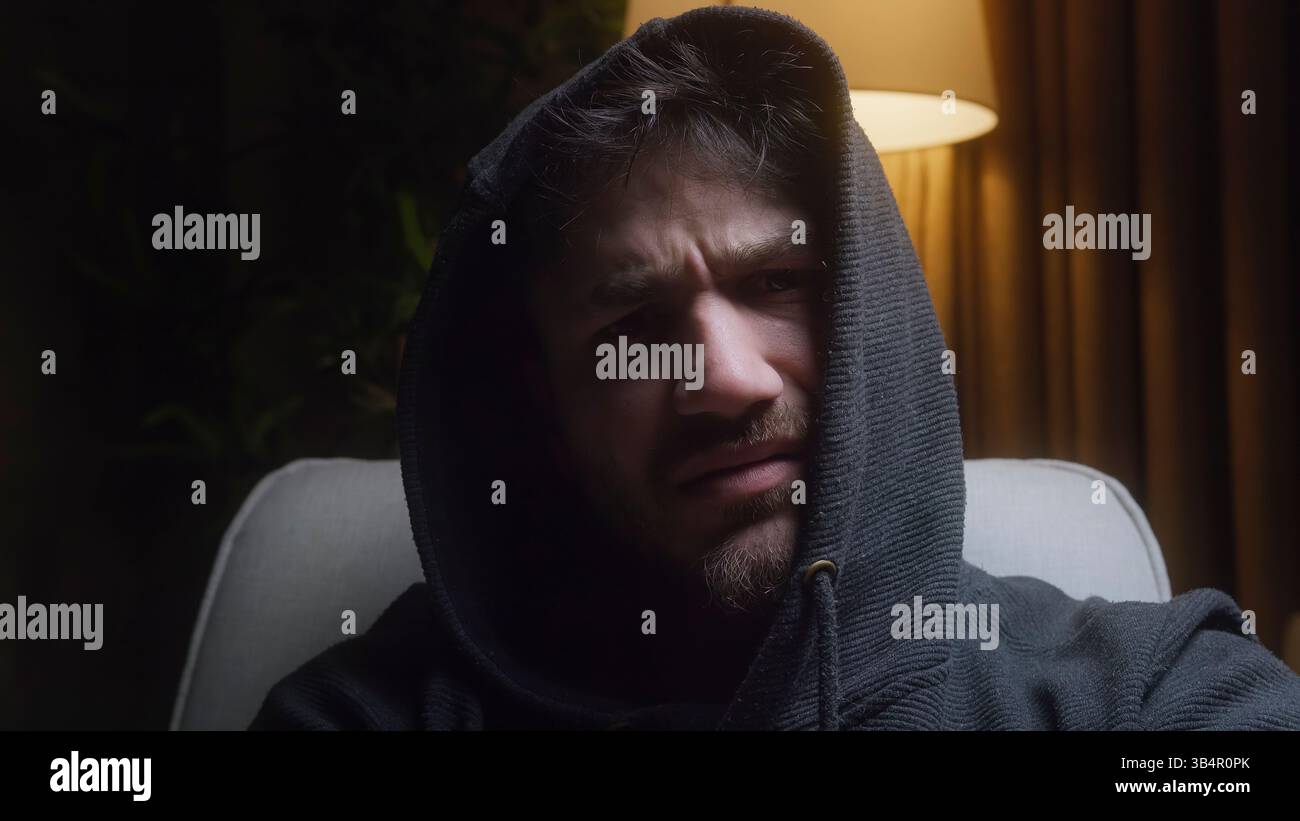 Verwirrter junger Mann, der einen Hoodie trägt, auf einer Couch in einem schwach beleuchteten Raum sitzt, die Stirn runzelt und einen seltsamen Blick gibt, während eine warme Stehlampe leise leuchtet Stockfoto