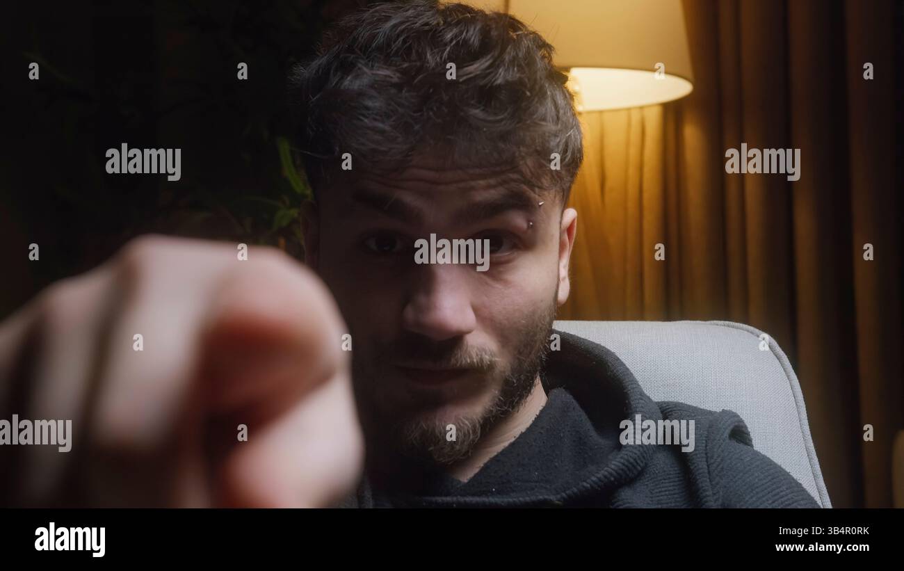 Junger Mann, der in schwach beleuchtetem Raum sitzt und auf die Kamera mit ernstem Ausdruck zeigt Stockfoto