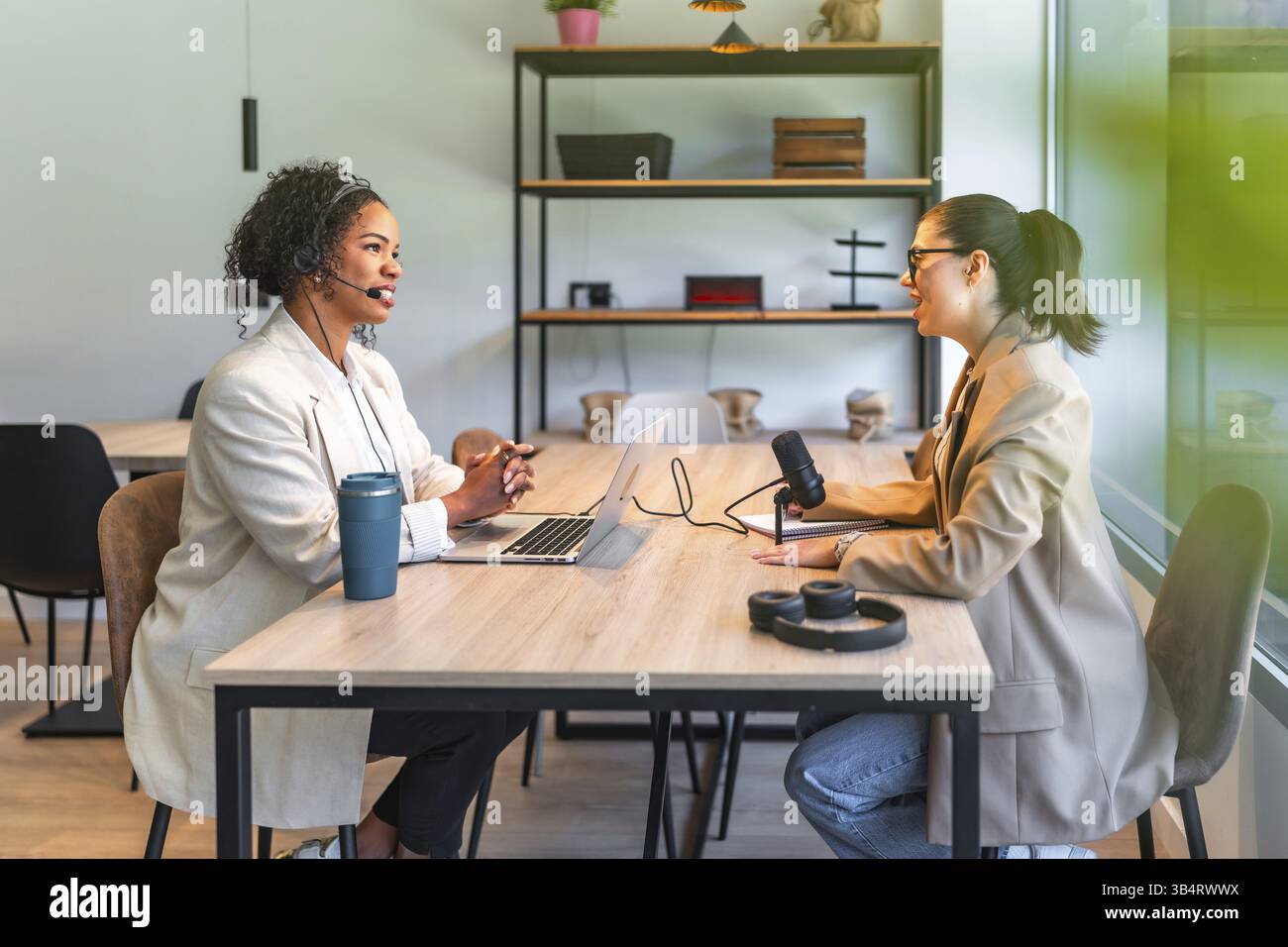 Zwei weibliche Podcasterinnen nehmen in einem modernen Büro eine neue Episode auf, die mit einem Mikrofon und einem Laptop ausgestattet ist Stockfoto