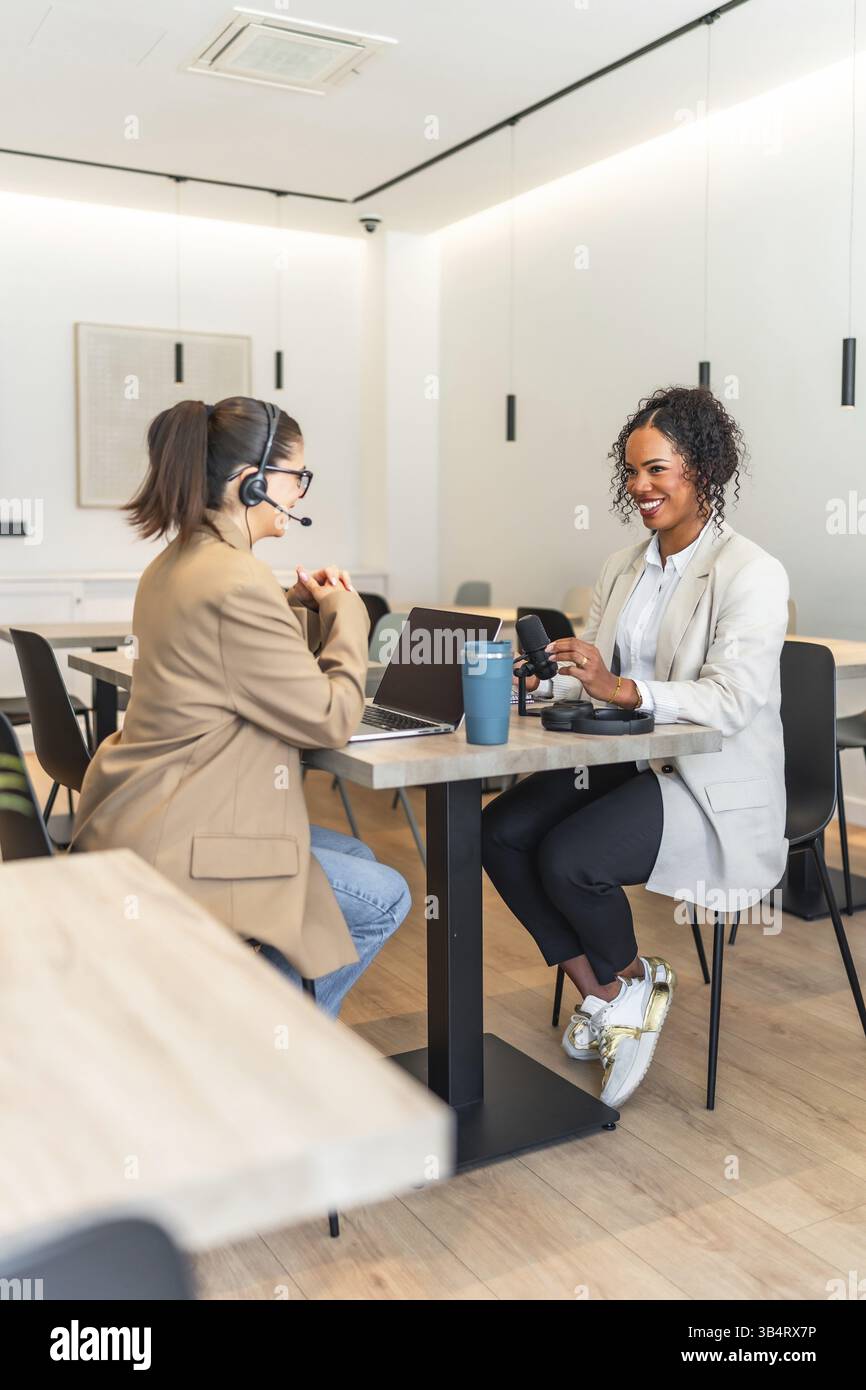 Zwei weibliche Podcasterinnen nehmen in einem modernen Büro eine neue Episode auf, die mit einem Mikrofon und einem Laptop ausgestattet ist Stockfoto