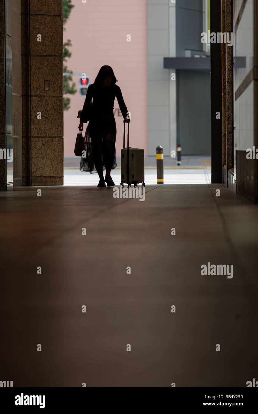 Silhouette einer Frau, die durch einen dunklen Korridor im Stadtgebäude geht, Koffer und Tasche zieht. Heller Hintergrund sorgt für einzigartigen Kontrast. Stockfoto
