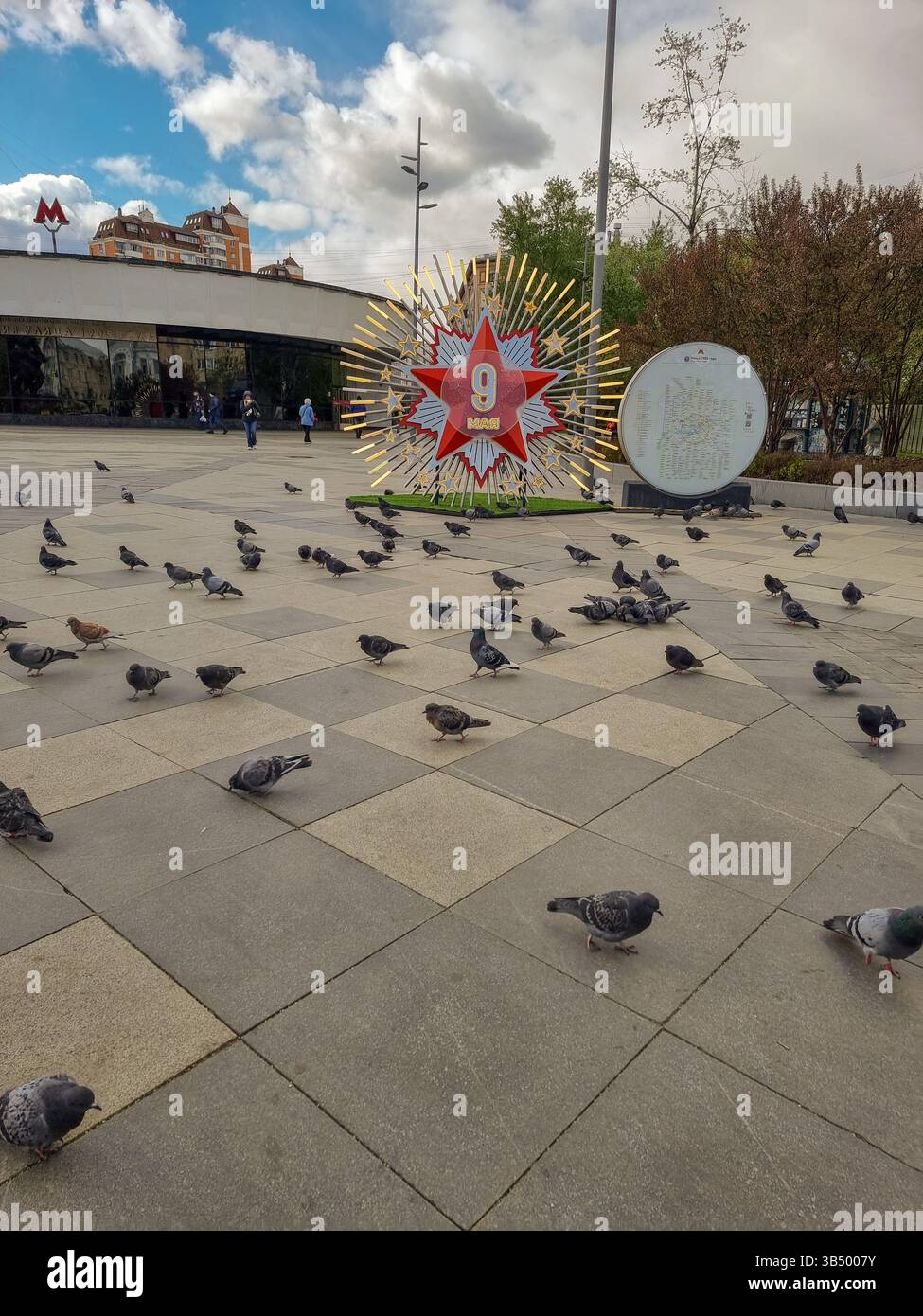 Tauben in der Nähe des Schildes für die Feier des 9. Mai 2025, 80 Jahre nach dem Sieg über die Nazis, Ende des 2. Weltkriegs: Leuchtender roter Stern mit 9. Mai auf russisch, auf Platz - Smartphone-aufgenommenes Stockfoto