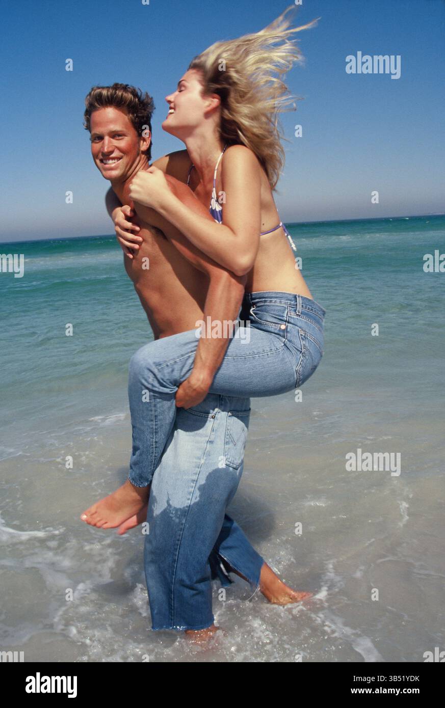 Lifestyle. Glückliches junges Paar in der Brandung. Mann, der eine Frau mit Huckepack trägt. Stockfoto