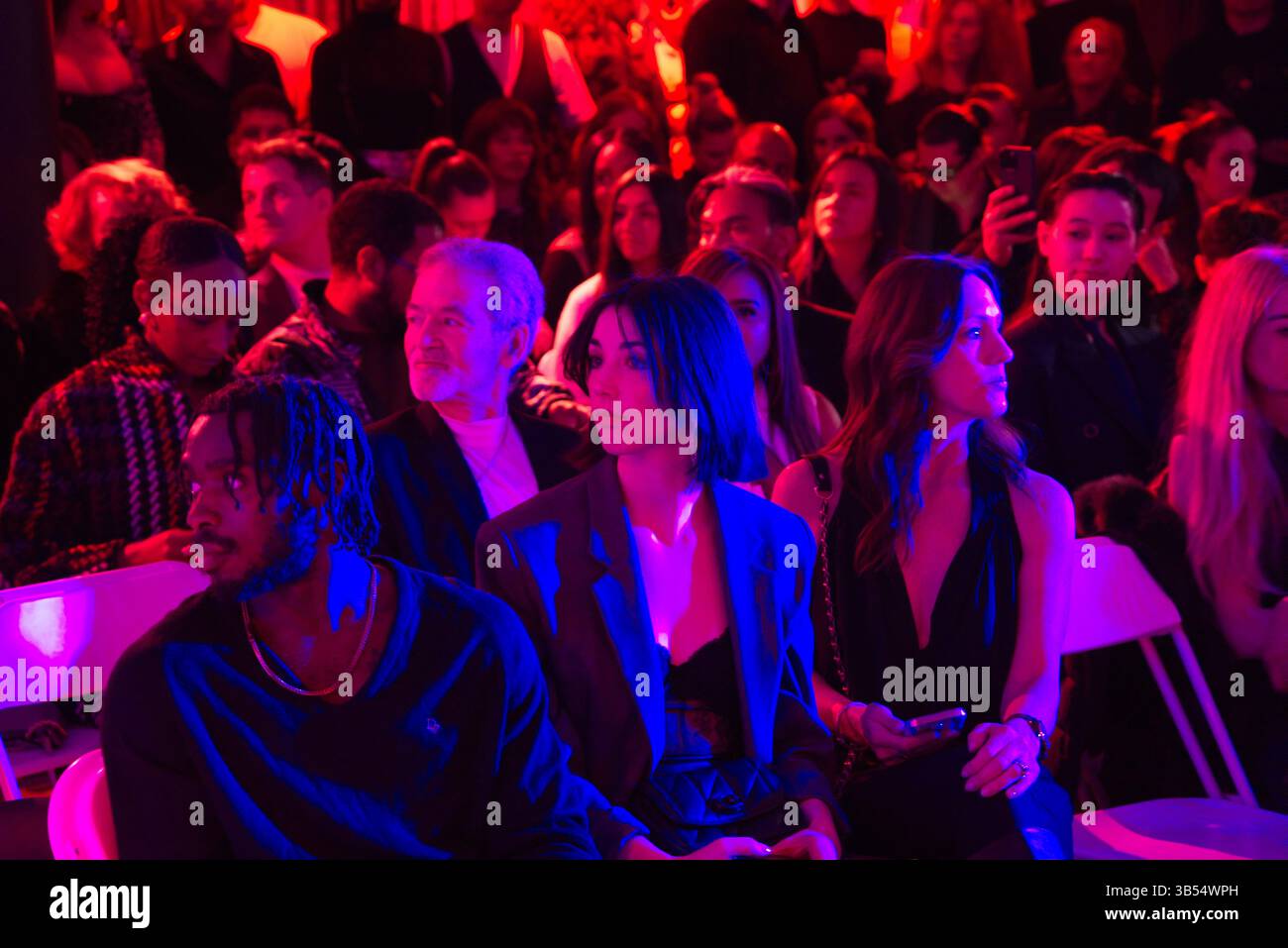 In einem schwach beleuchteten Raum unter violetter und blauer Beleuchtung sitzt eine vielfältige Gruppe von Menschen, überwiegend Frauen. Sie scheinen auf Mode zu gehen Stockfoto