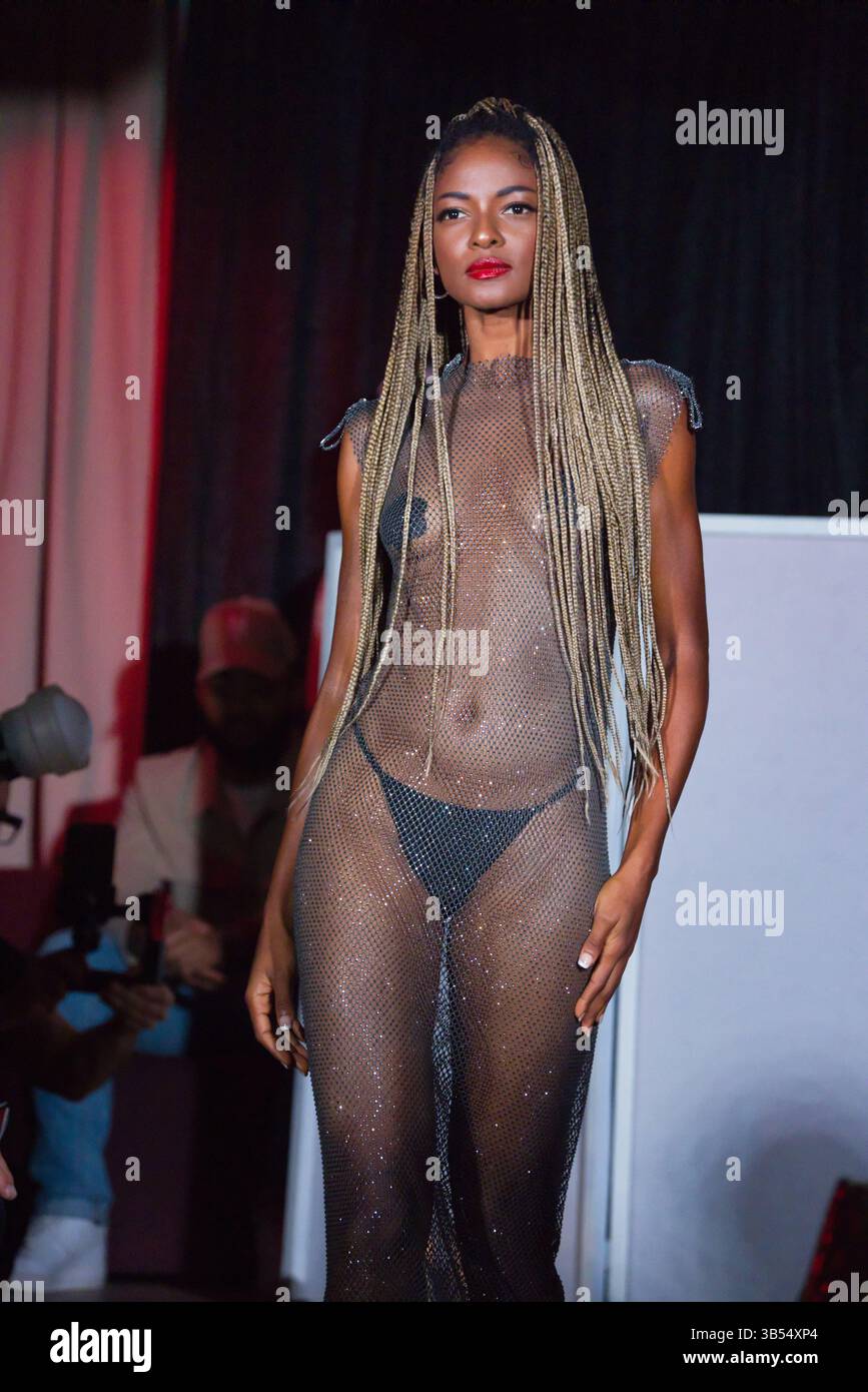 Eine dunkelhäutige Frau mit langen blonden Zöpfen modelliert ein transparentes, dunkles, glitzerndes Maxi-Kleid aus Mesh mit einem schwarzen Bikiniboden darunter. Sie steht Stockfoto