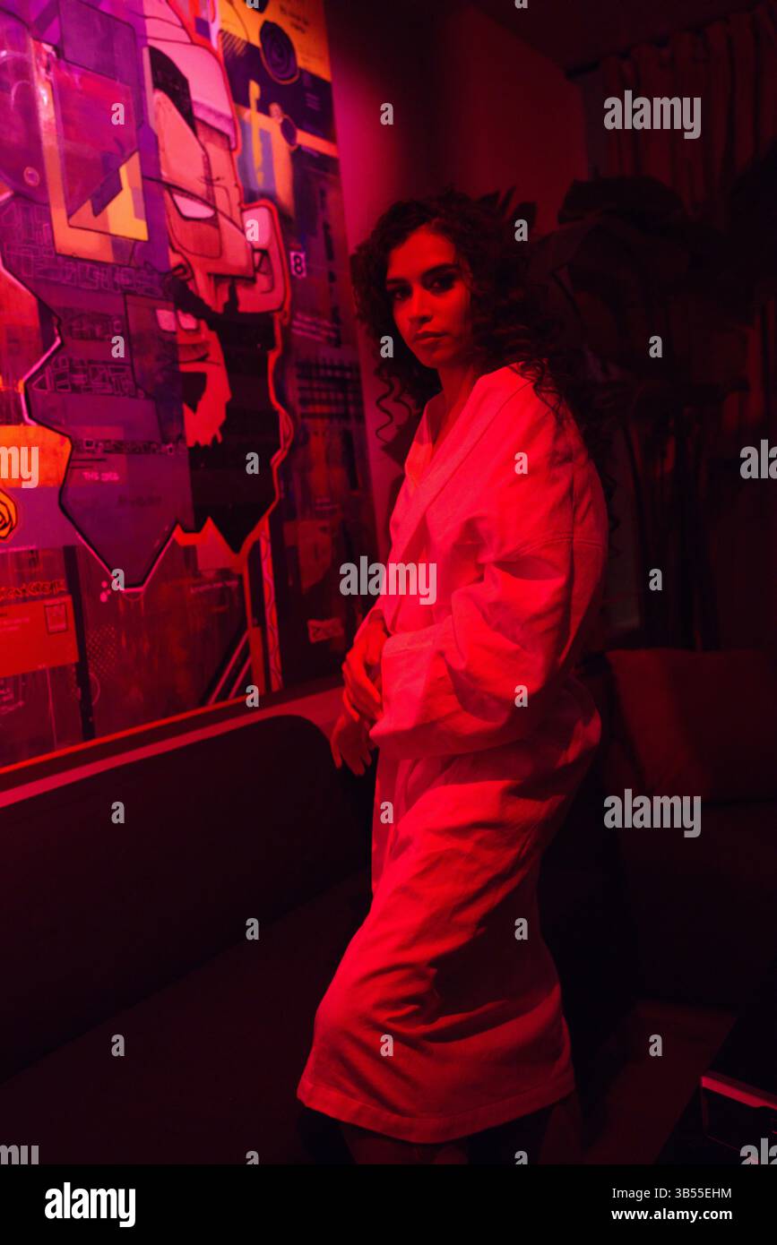 Eine Frau mit lockigen braunen Haaren steht in einem schwach beleuchteten Raum, der in rotes Licht getaucht ist. Sie trägt ein langes, helles Gewand. Eine große, bunte abstrakte Malerei Stockfoto