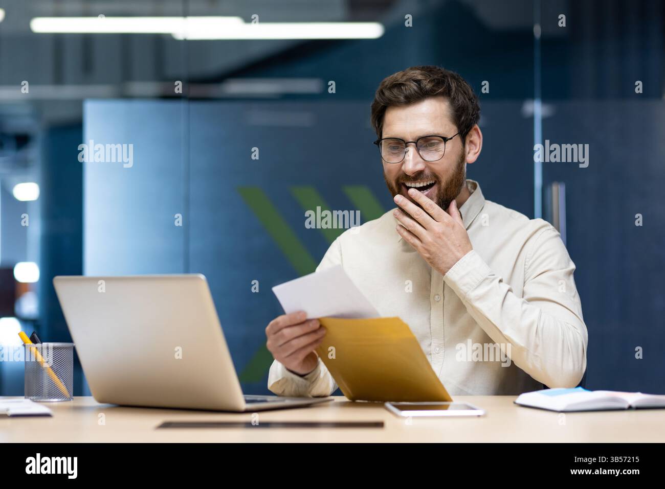 Ein glücklicher junger Mann sitzt im Büro an einem Laptop und liest einen erhaltenen Brief, freut sich über die gute Nachricht. Stockfoto
