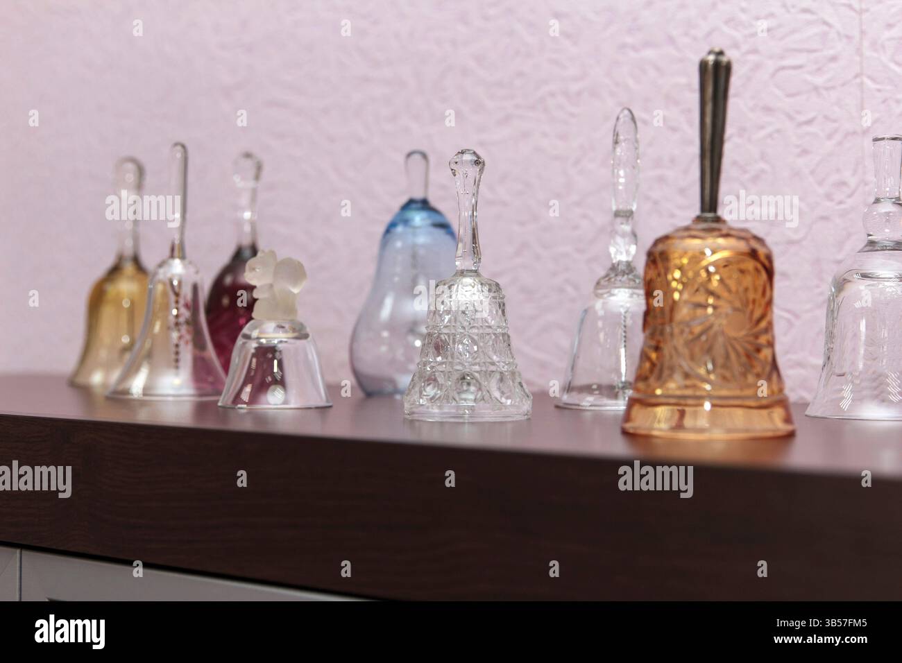 Verschiedene transparente und farbige Glasglocken, die auf einem dunklen Holzregal vor einer rosafarbenen strukturierten Wand angeordnet sind, zeigen eine Kollektion von Wohnkultur Stockfoto