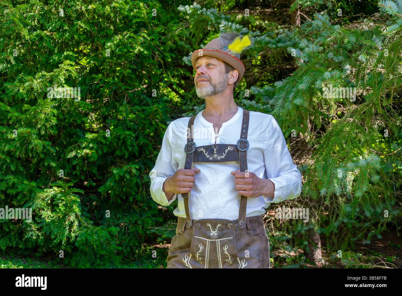 Ein Mann in traditioneller bayerischer Kleidung posiert vor einem Hintergrund grüner Bäume Stockfoto