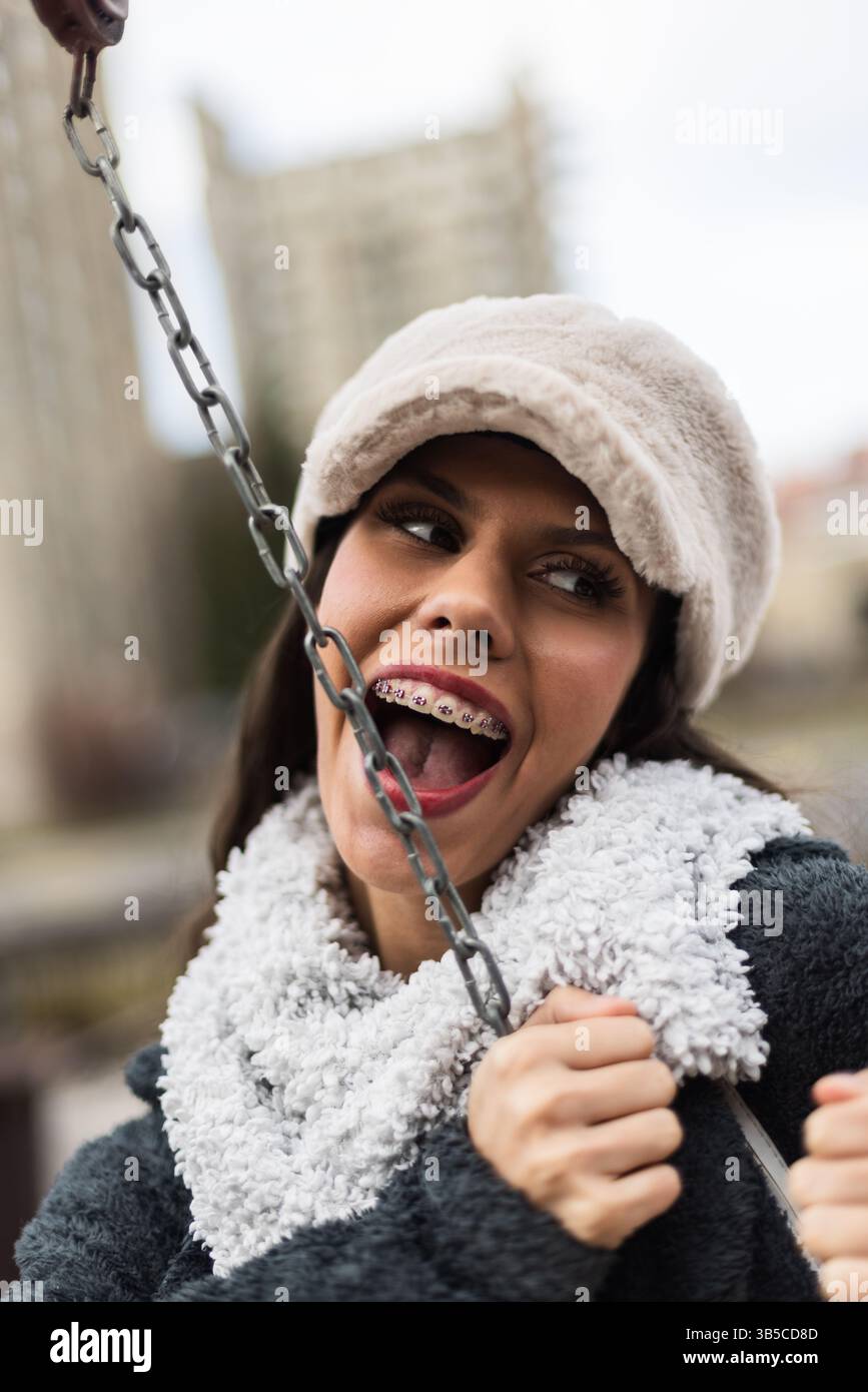 Eine fröhliche junge Frau mit Hut und Schal lächelt draußen und zeigt einen fröhlichen Ausdruck und eine Zahnspange. Sie genießt eine lustige Aktivität gegen Stockfoto