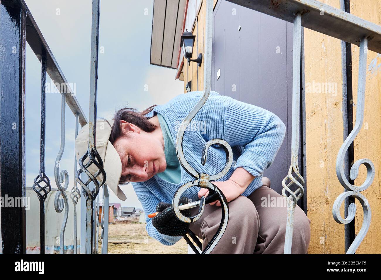 Mit präzisen Pinselstrichen, um verwitterte Veranden zu beschichten, belebt reifes Individuum sorgfältig kunstvoll verzierte Metallrahmen neben dem Eingang. Stockfoto