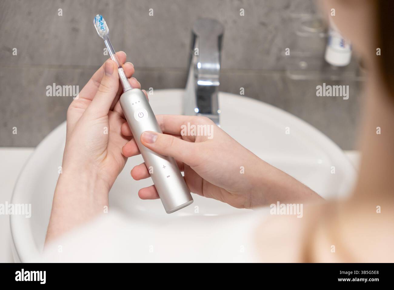 Die Hände einer Frau halten den Griff der elektrischen Zahnbürste und den abnehmbaren Bürstenkopf vorsichtig über ein Waschbecken, um die wichtigsten Vorbereitungsschritte für zu veranschaulichen Stockfoto